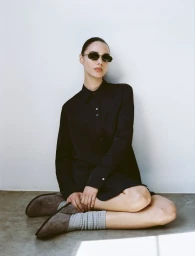 Model wearing a dark buttoned shirt dress and grey socks sitting against a white wall.