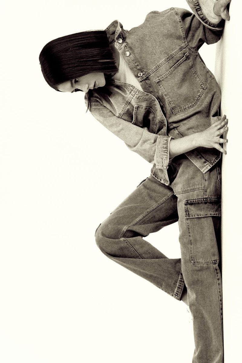 Monochromatic portrait of a woman in a denim jacket and pants leaning against a white wall with subtle shadows. Fashion editorial.