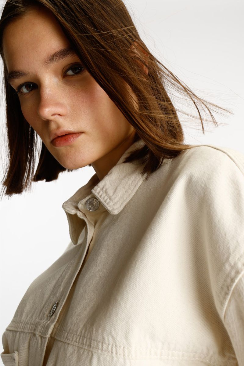Close-up portrait of a woman with a brown bob wearing an off-white denim jacket against a clean white backdrop.