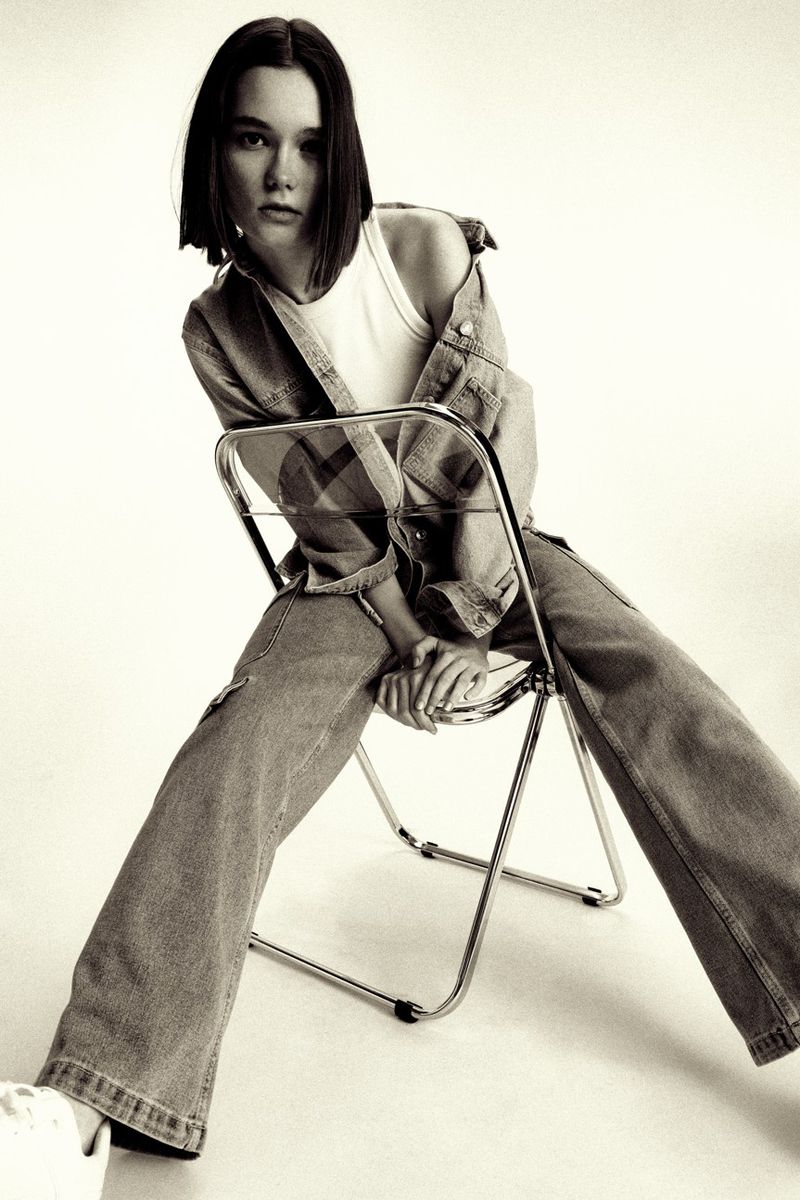 Model with short dark bob hair seated on a folding chair wearing a denim jacket, wide-leg jeans, and white tank top.