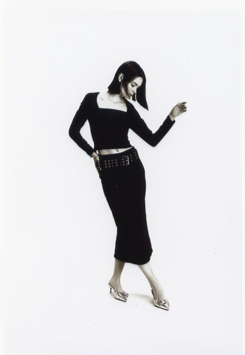 A woman in a black long-sleeved crop top and skirt poses against a white backdrop, accented by a studded belt and pointed silver heels.