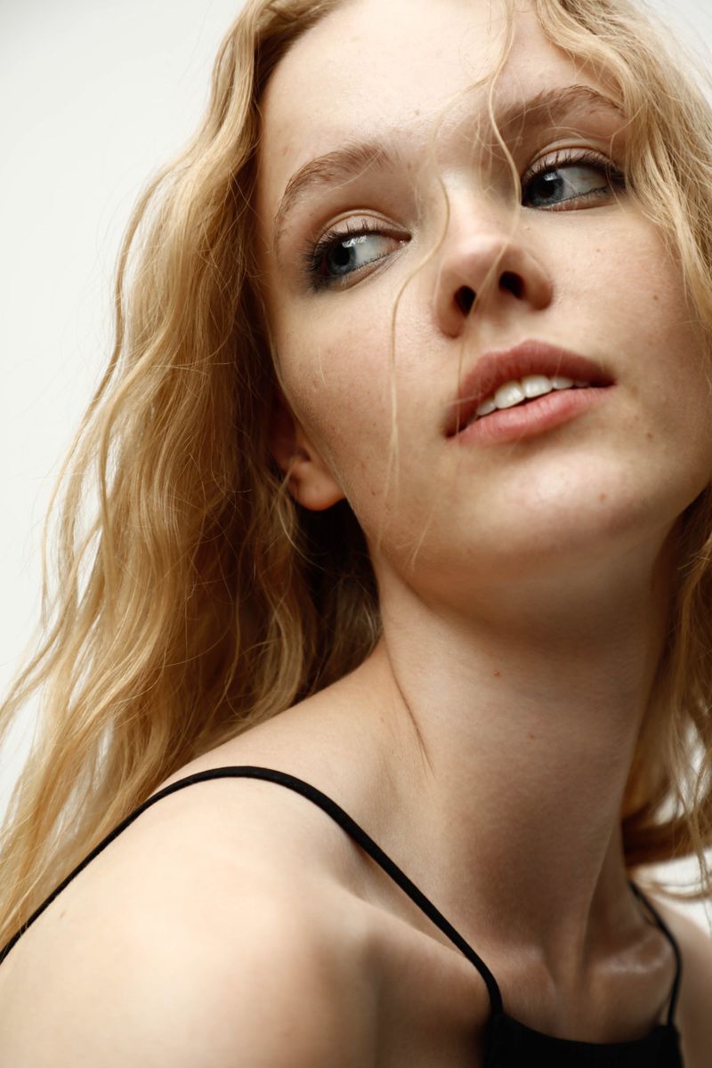 Headshot portrait of a young adult female model with blonde wavy hair wearing a black top against a neutral backdrop, looking away.