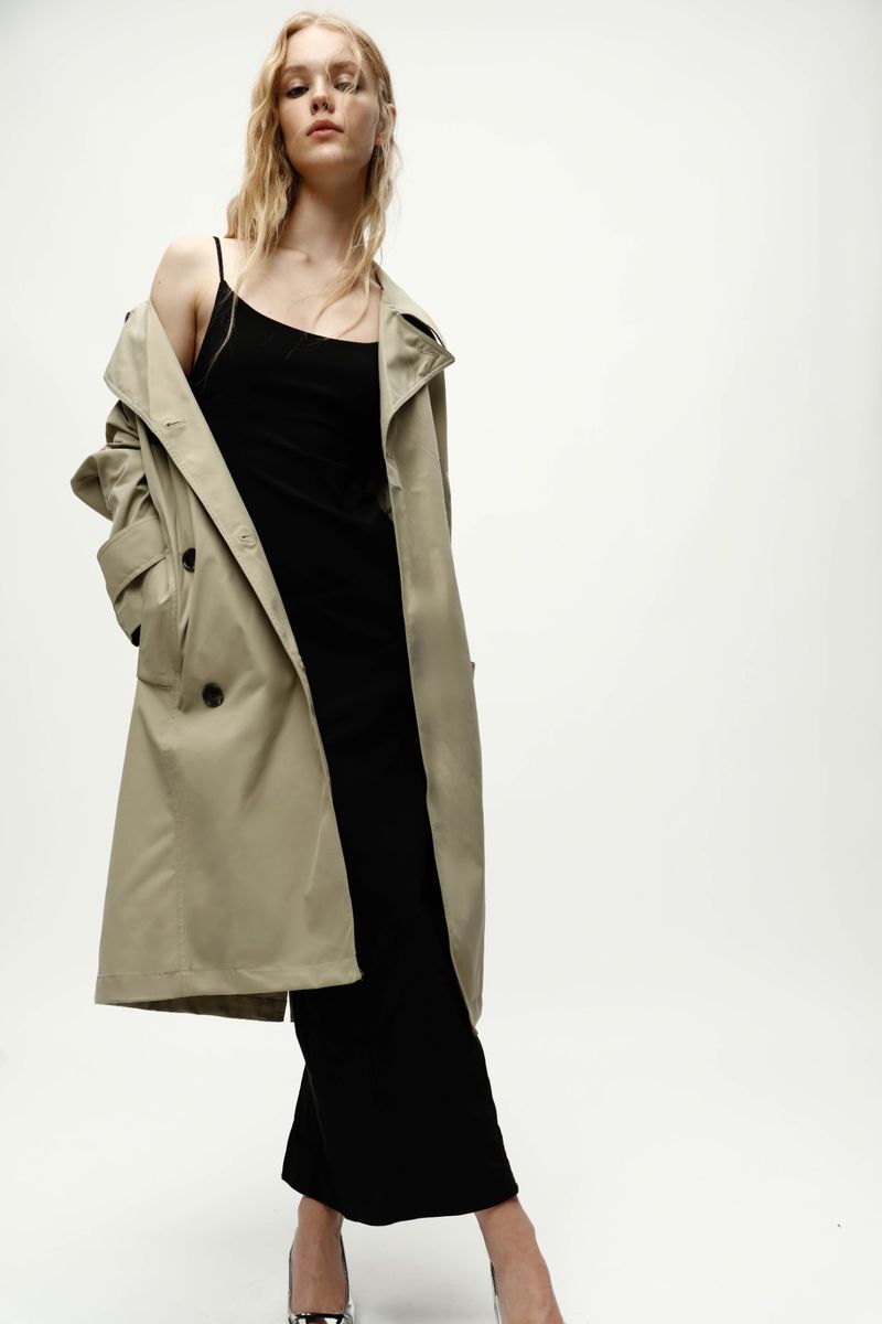 A fair-skinned woman with blonde hair is posing in a black dress and beige trench coat against a white background in a fashion shot.