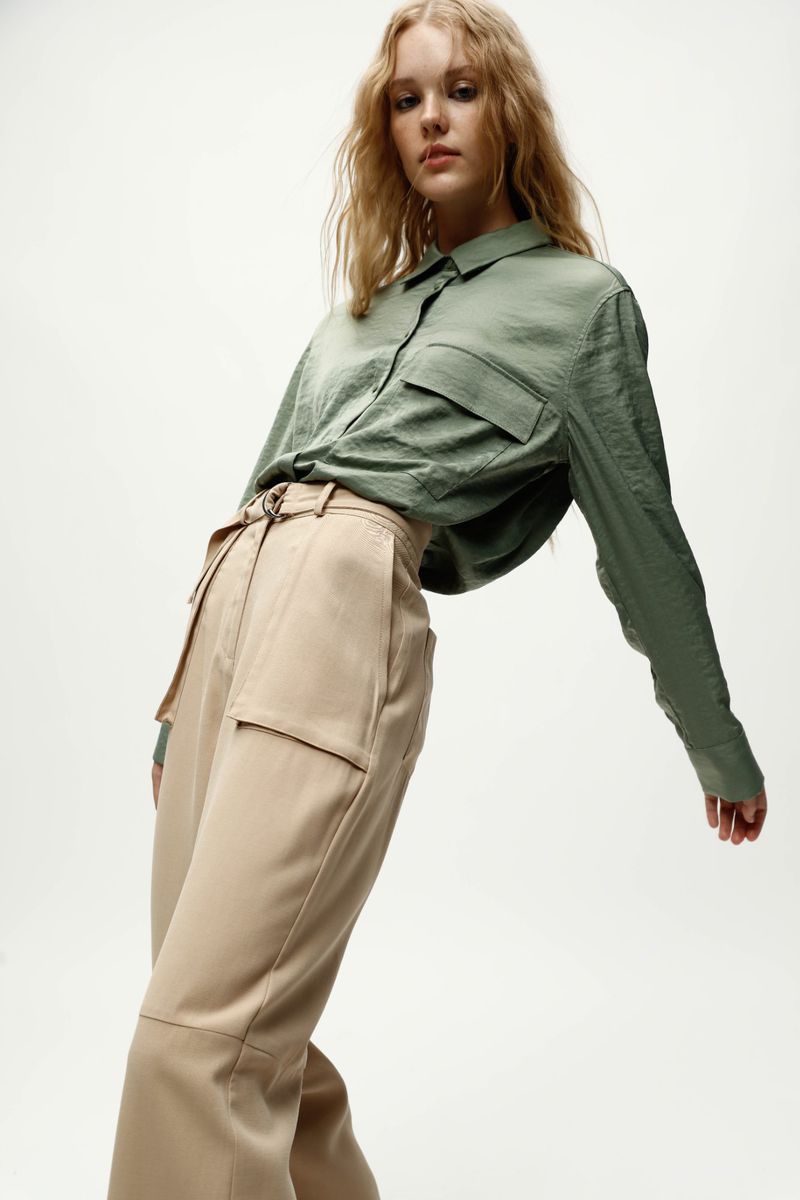 Fashionable woman poses in a stylish green button-down shirt and wide, high-waisted tan pants against a neutral backdrop in a studio shot.