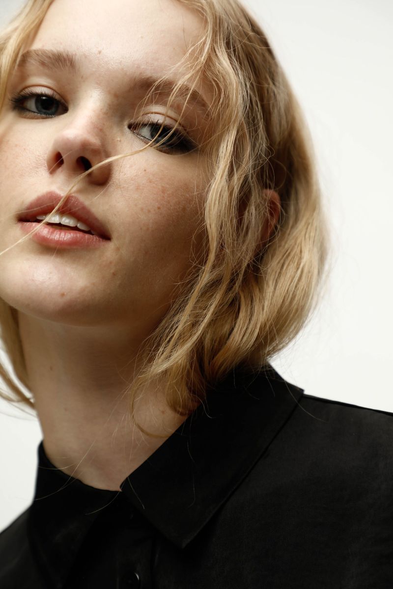 Close-up portrait of a blonde woman wearing a black collared shirt against a white seamless background.