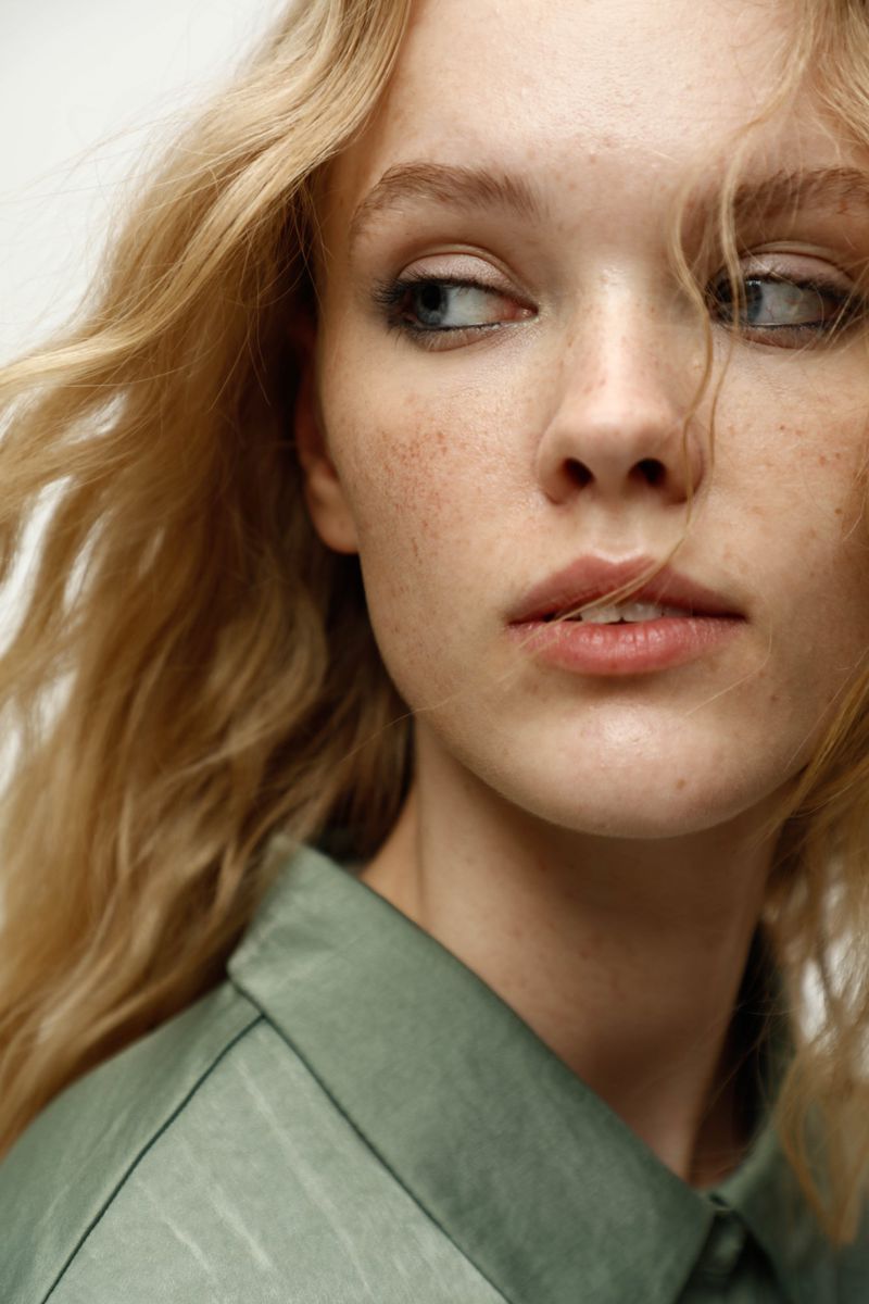 Close-up portrait of a young woman with wavy blonde hair and light skin, wearing a green collared shirt against a white background.