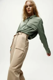 Model with long blonde hair wearing a green button-up shirt and high-waisted beige trousers against a white background