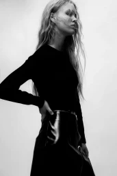 Monochrome fashion editorial portrait of a woman wearing a black long-sleeve top and glossy dark skirt against a pale background.
