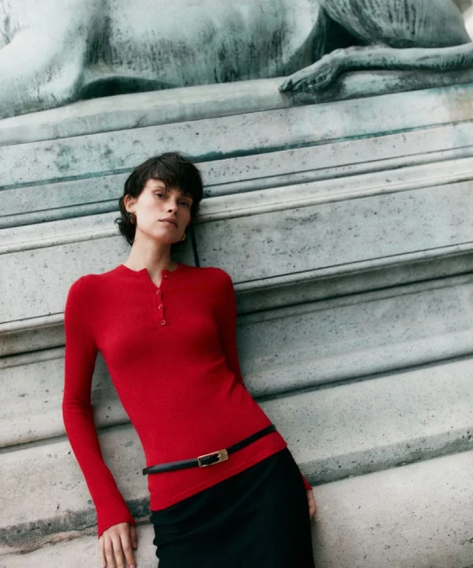 Elegant adult woman in red long-sleeve shirt and black skirt leans against a stone wall featuring a relief sculpture.