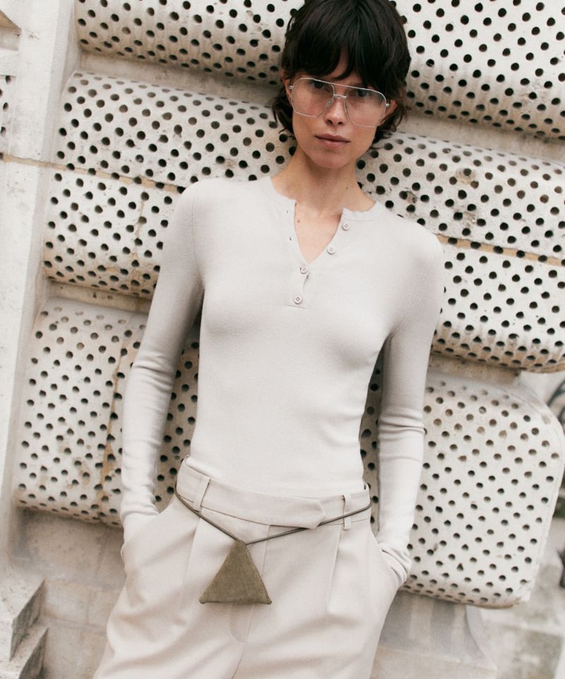 A light-skinned woman with short dark hair models a pale-grey, long-sleeve top and trousers in front of a textured white background. 