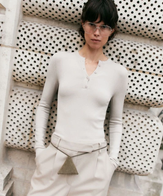 A light-skinned woman with short dark hair models a pale-grey, long-sleeve top and trousers in front of a textured white background. 