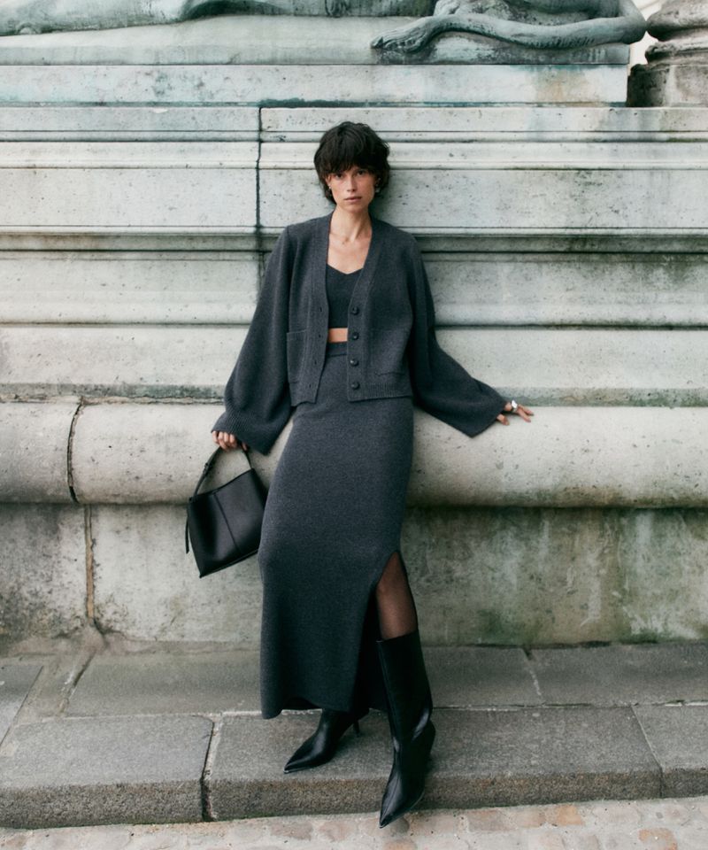 Woman in a gray knit skirt suit and black boots poses against a stone wall, holding a black leather purse for a fashion shoot.