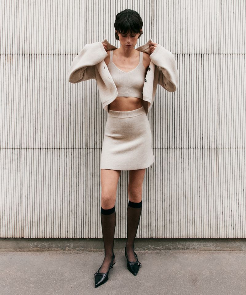 Woman in a knitted crop top, cardigan and skirt poses against a textured wall, accessorized with knee-high socks and pointed-toe flats.