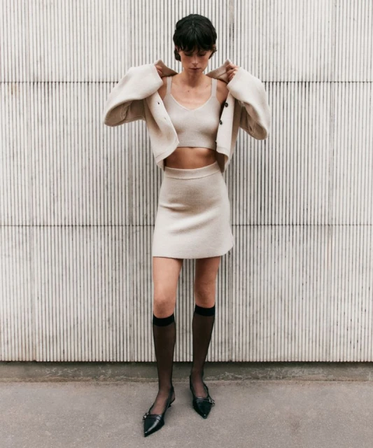 Woman in a knitted crop top, cardigan and skirt poses against a textured wall, accessorized with knee-high socks and pointed-toe flats.