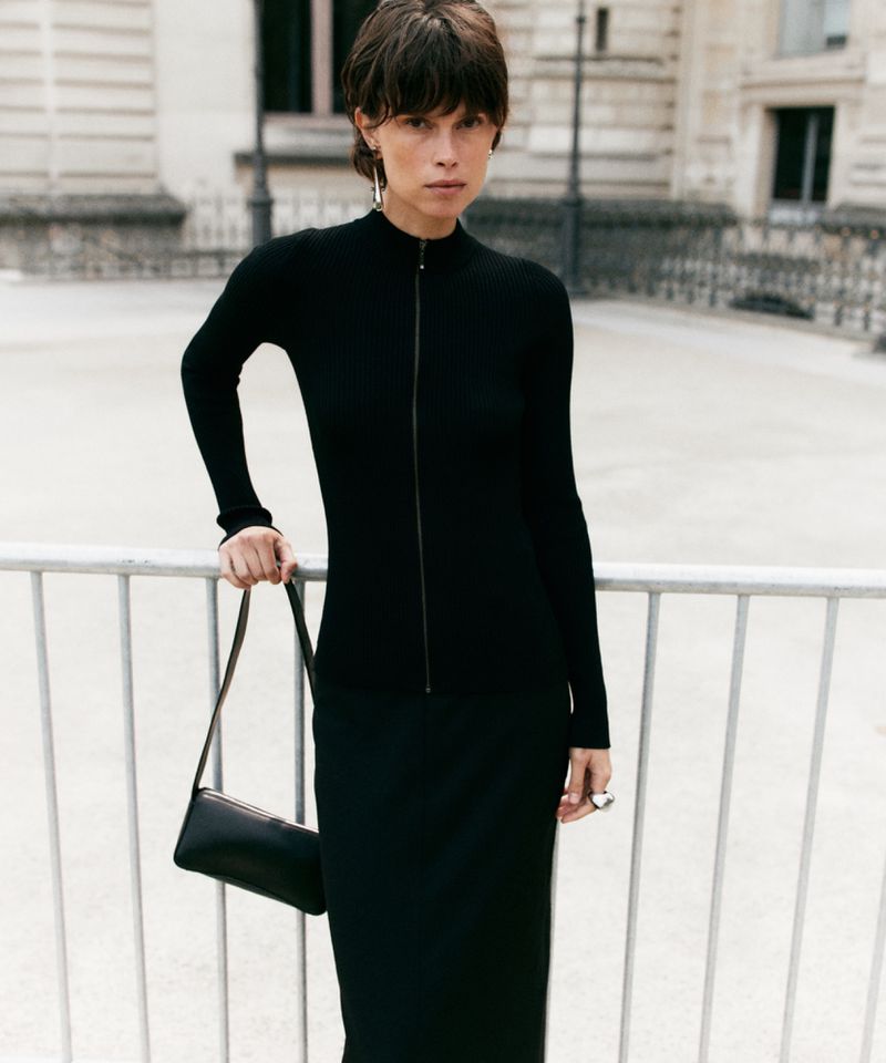 Woman in a black zip-up long-sleeve dress stands outside with a black bag. The background shows a light-colored building with architectural detail.