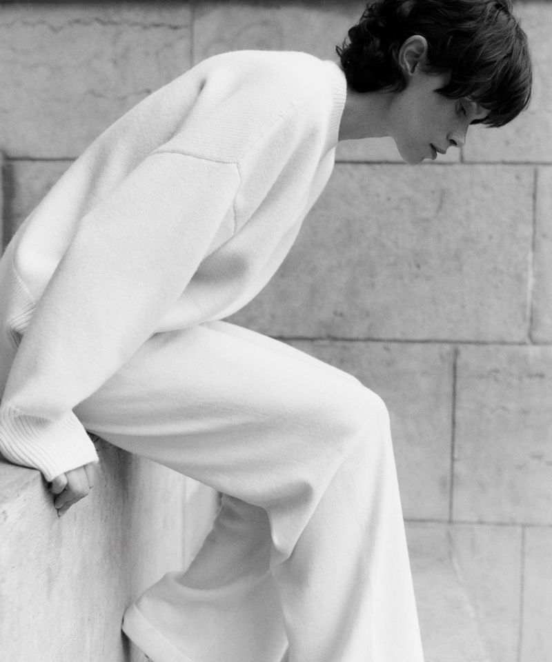 Black and white portrait of a young adult wearing a matching sweater and wide-leg pants, posing against a stone wall backdrop.