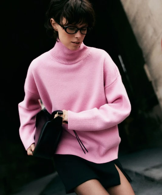 A stylish woman wearing a pink sweater and a black skirt is holding a black bag, creating a fashionable and modern composition.