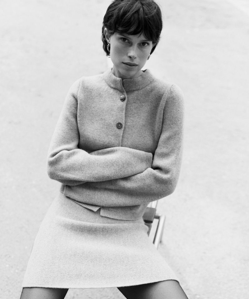 Monochrome portrait of a woman with short dark hair in a grey knit sweater and skirt, seated with arms crossed in a simple outdoor setting.
