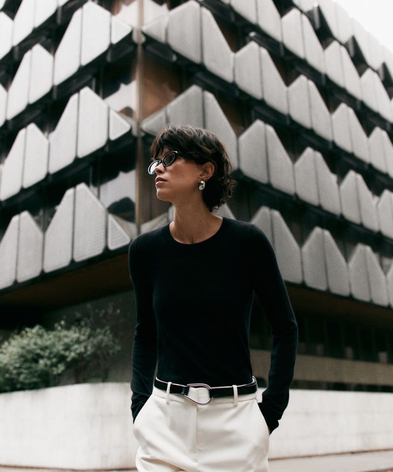 Adult woman in a black long-sleeve shirt and white pants poses outdoors with a building featuring a geometric facade in the background.