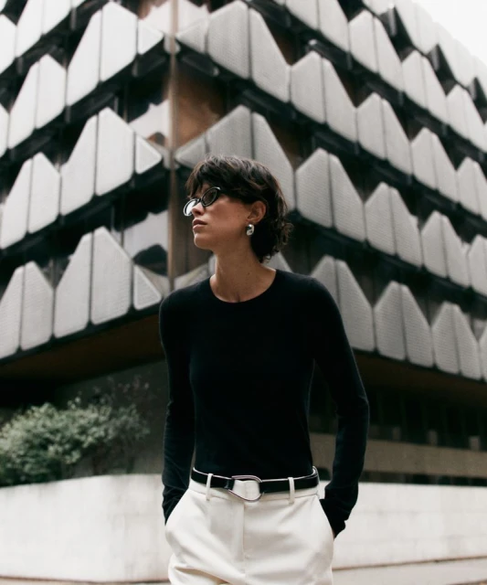 Adult woman in a black long-sleeve shirt and white pants poses outdoors with a building featuring a geometric facade in the background.