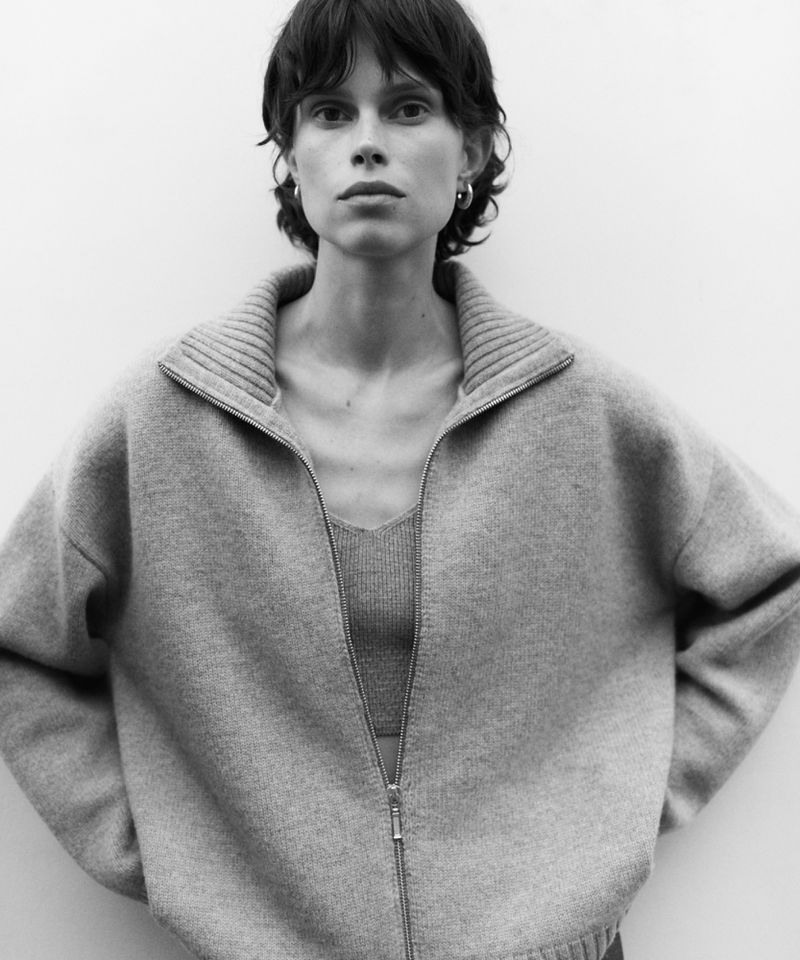 Monochrome portrait of a woman with short curly hair wearing a wool zip-up sweater against a neutral white background in a studio.