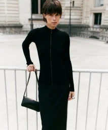 Model in black zip-up top and matching skirt holding a black handbag outdoors