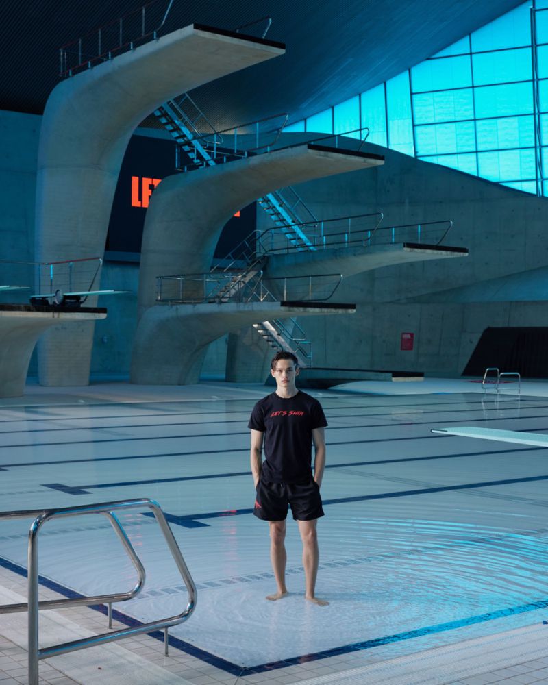 Indoor shot of a lifeguard standing in shallow water in a modern swimming pool complex with diving boards and bright blue accents.