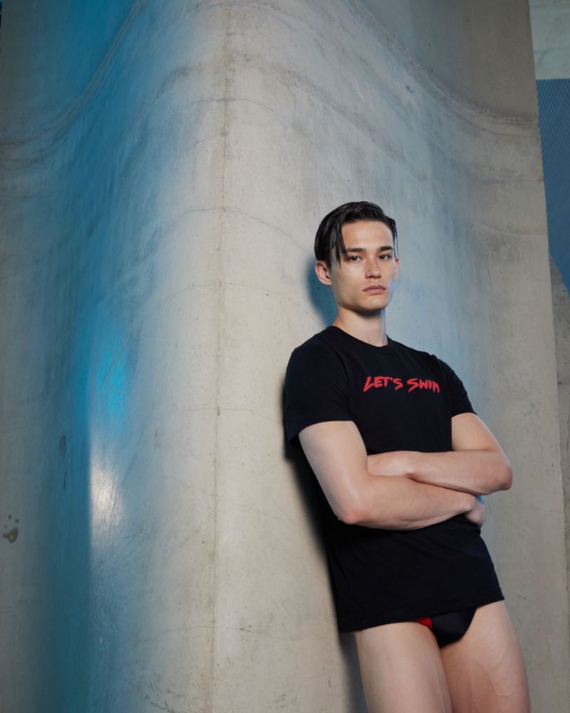 A young man in a black t-shirt poses against a concrete wall with blue lighting, creating a simple yet striking studio portrait.