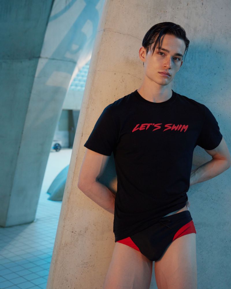 Athletic man poses in swimwear and t-shirt against architectural concrete. Black swimwear, red accents, modern architecture, indoor pool setting.