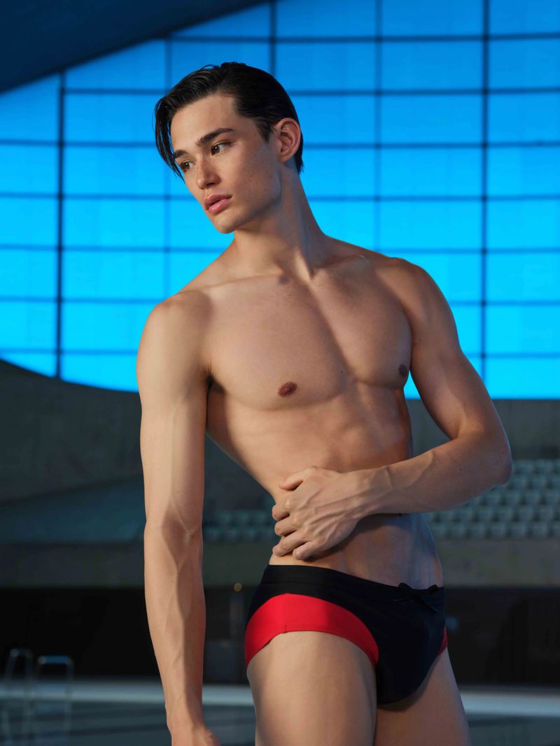 A muscular adult male poses shirtless in swim trunks in a medium shot against a blurred light blue background in an indoor setting.