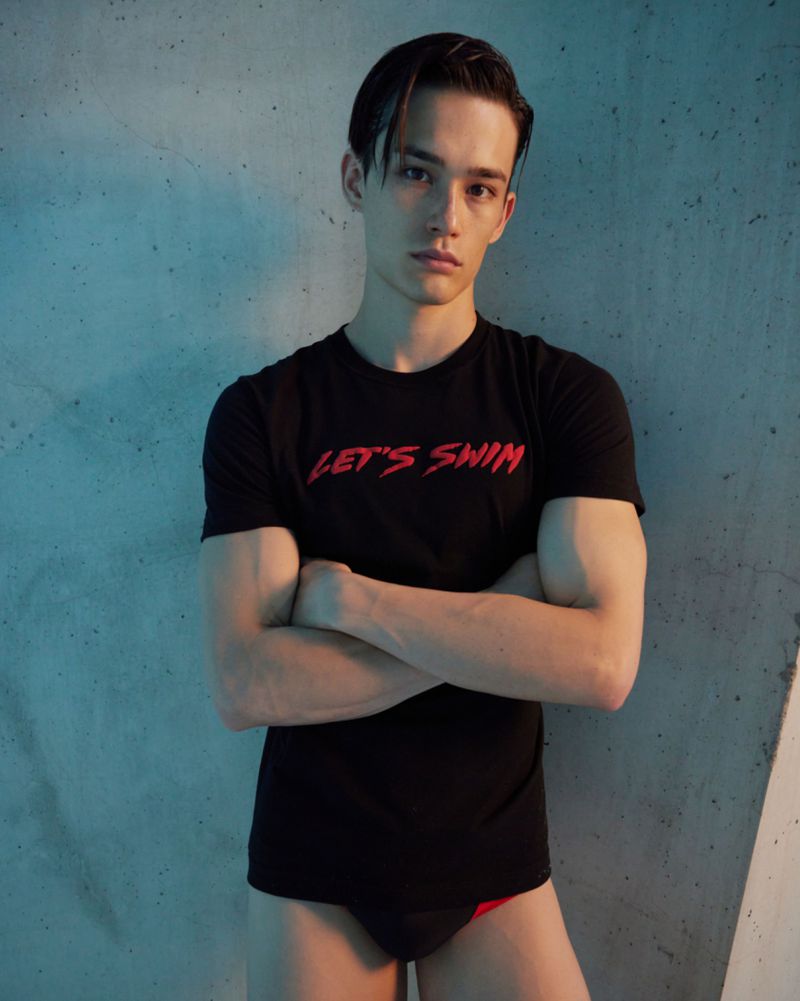 Portrait of a young adult male in a black t-shirt, arms crossed, against a textured gray wall with blue gel lighting on one side.