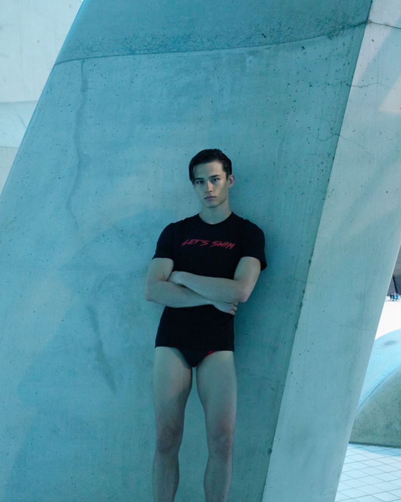 Young man in swimwear poses by textured blue wall; he wears a t-shirt that says 'Let's Swim'. Indoor setting with cool lighting.