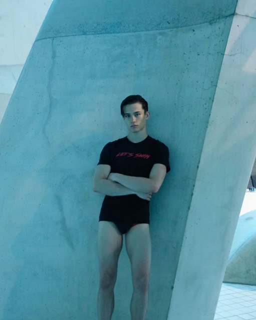 Young man in swimwear poses by textured blue wall; he wears a t-shirt that says 'Let's Swim'. Indoor setting with cool lighting.