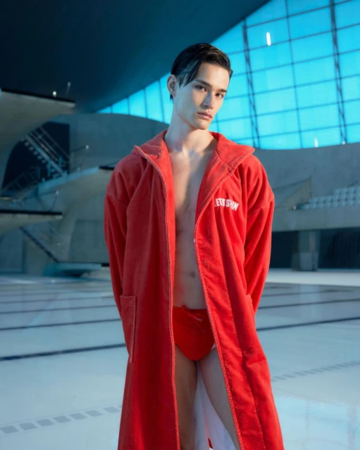 Young man poses in red swimsuit and robe by swimming pool with natural light and modern architecture, showcasing fitness and fashion.