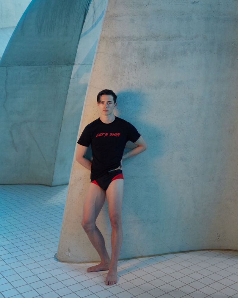 Athletic man poses in a black t-shirt and swimwear against a blue-toned architectural backdrop, standing barefoot on tiled flooring.
