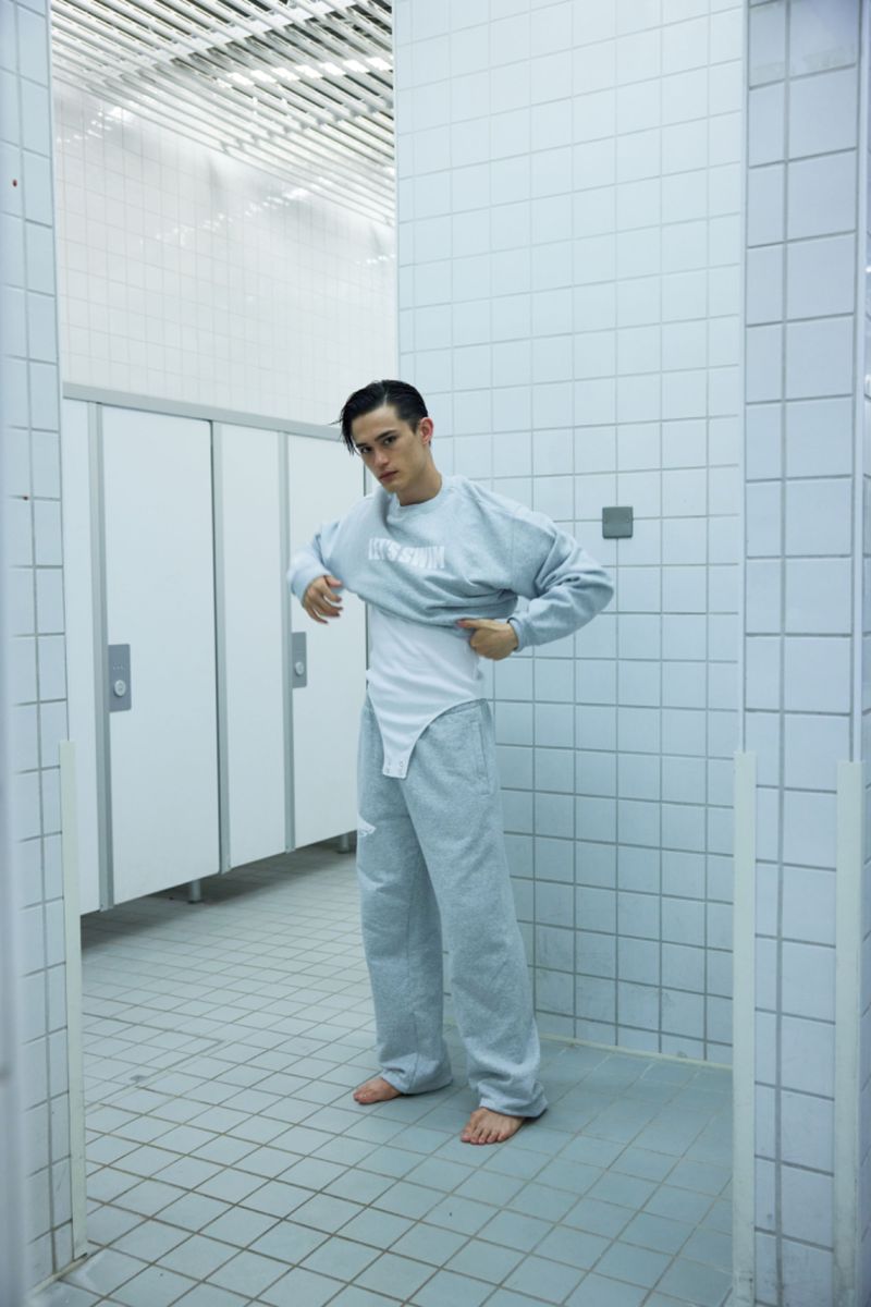 Young adult male in a public restroom adjusting a light grey sweatshirt and matching sweatpants, wearing a white bodysuit underneath.