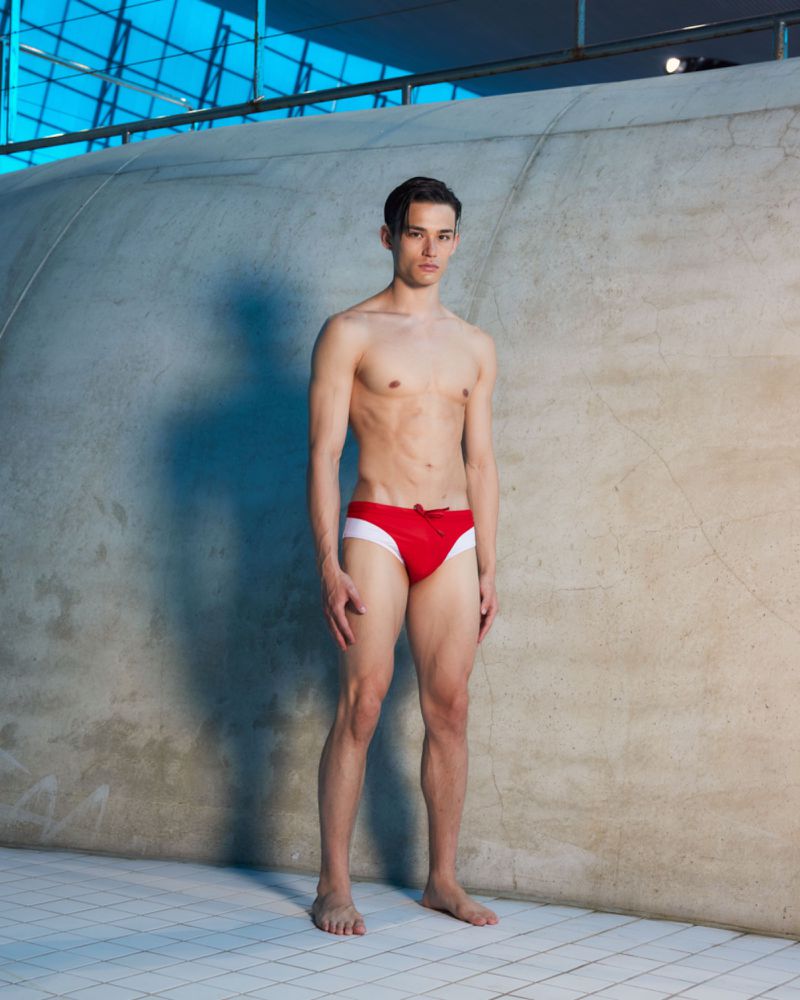 A slender adult male poses indoors wearing a red swimsuit against a curved wall and tile floor, bathed in soft, diffused lighting.