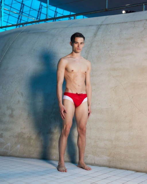 A slender adult male poses indoors wearing a red swimsuit against a curved wall and tile floor, bathed in soft, diffused lighting.