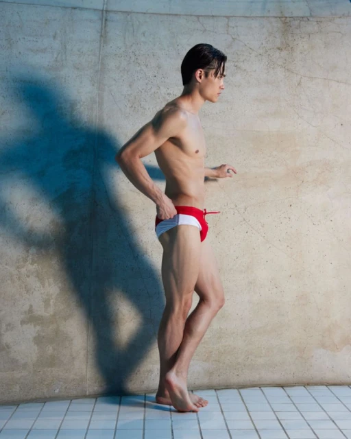 Athletic man stands sideways in red and white swimwear next to a concrete wall, with a shadow cast on the wall from side lighting.