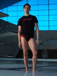 Model Alex Schlab standing in shallow water wearing a black Lets Swim t-shirt and swim briefs by an indoor pool.