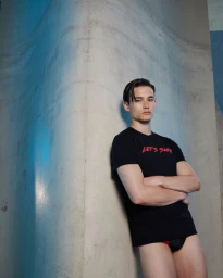 Alex Schlab wears a black Lets Swim graphic t-shirt and swim briefs while leaning against a curved concrete wall.