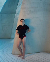 Alex Schlab standing barefoot in a black t-shirt and red-trimmed swimwear against a curved concrete wall.