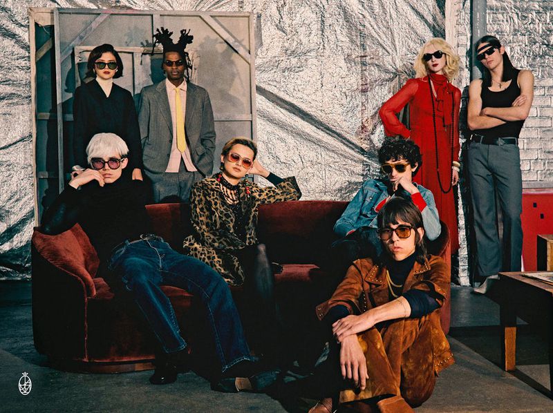 Group of eight people indoors. The people are wearing retro style clothing and sunglasses, sitting and standing in front of a silver background.