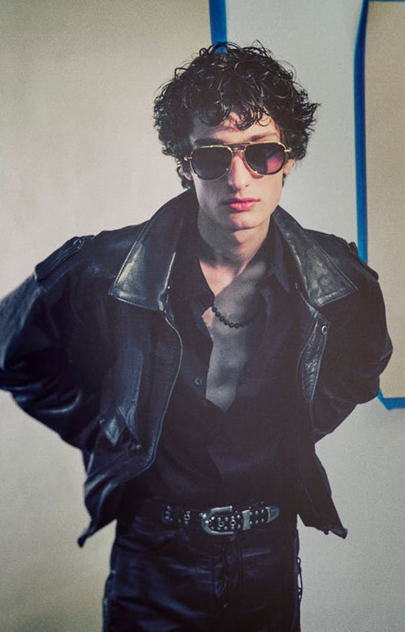 Stylish man in sunglasses and leather jacket strikes a casual pose against a neutral backdrop with blue accents, fashion shoot.