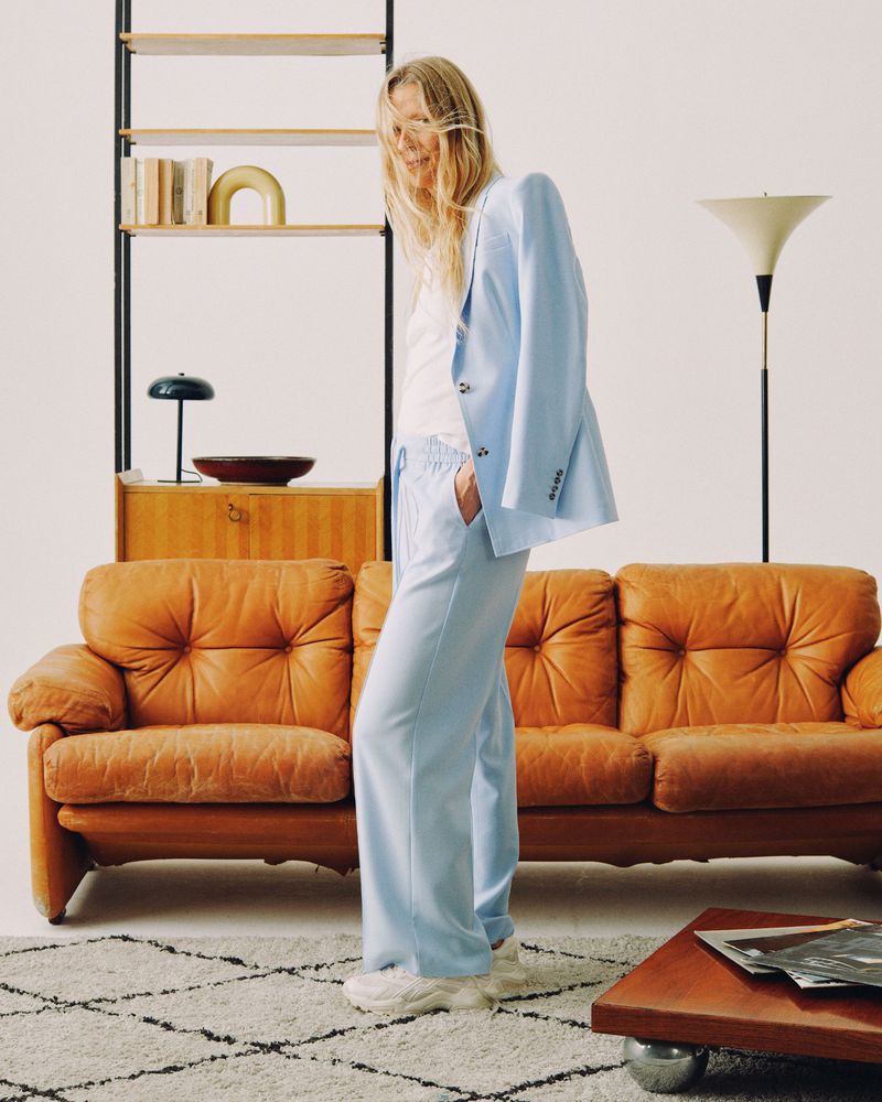 A woman in a light blue suit stands in a neutral indoor space, posing near a brown couch, with shelves and a geometric rug visible.