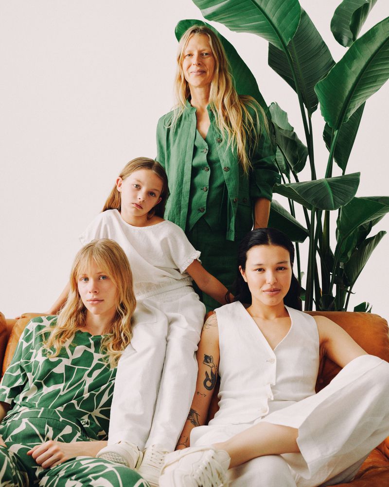 A group of four women, including a child, are casually posed on a brown couch in a studio with a large green plant in the background.