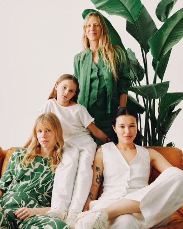 A group of four women, including a child, are casually posed on a brown couch in a studio with a large green plant in the background.