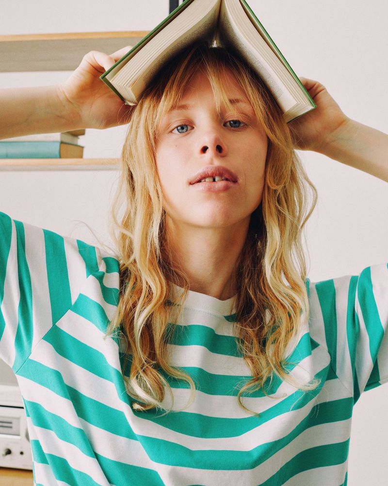 Young adult with blond wavy hair poses with a book balanced on their head, wearing a white and green striped shirt, indoors.