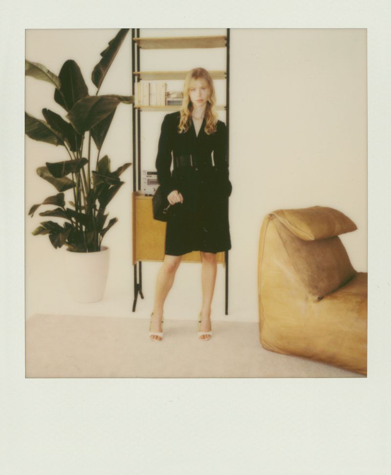 Fashion shot of a young adult woman posing in a short, belted black coat with high-heeled shoes, indoor setting with plant and chair.