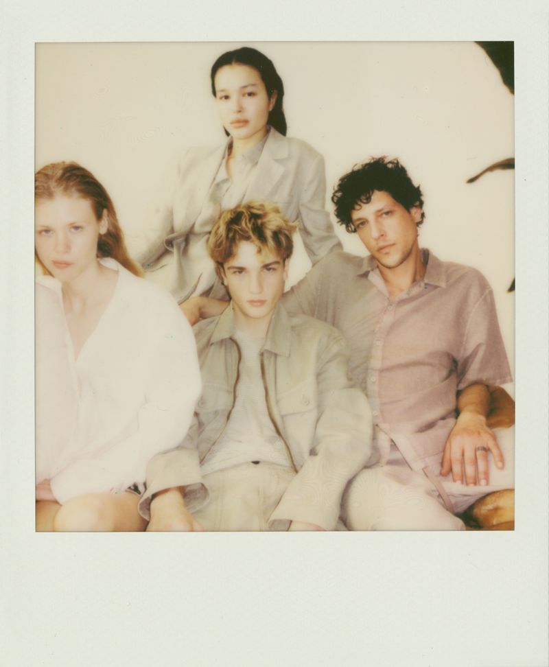 Four young adults in neutral-toned outfits pose for a polaroid-style portrait in a bright, casual studio setting with soft, diffused lighting.